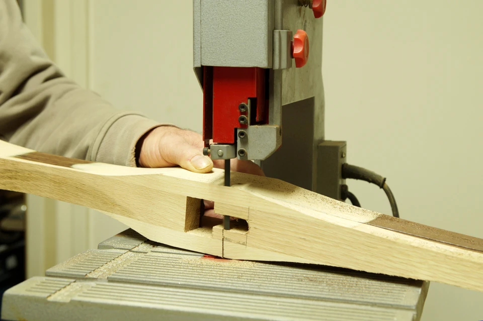 Atelier de menuiserie intérieur où un artisan en pull beige utilise une mortaiseuse à bédane pour percer des mortaises dans une pièce de bois massif