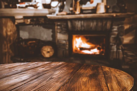 Intérieur rustique avec table en bois massif au premier plan, cheminée en pierre avec feu allumé en arrière-plan